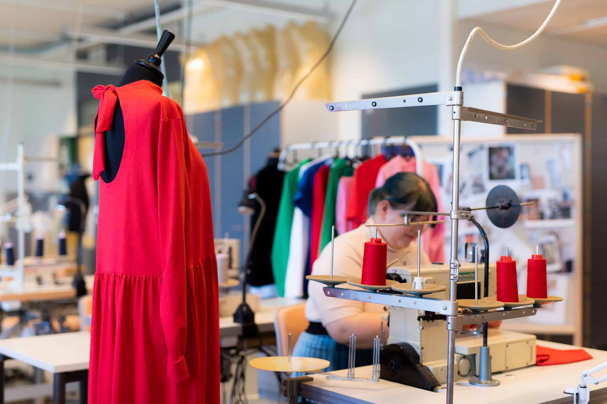 Etualalla punainen mekko sovitusnuken päällä. Taustalla nainen ompelee ompelukoneella.