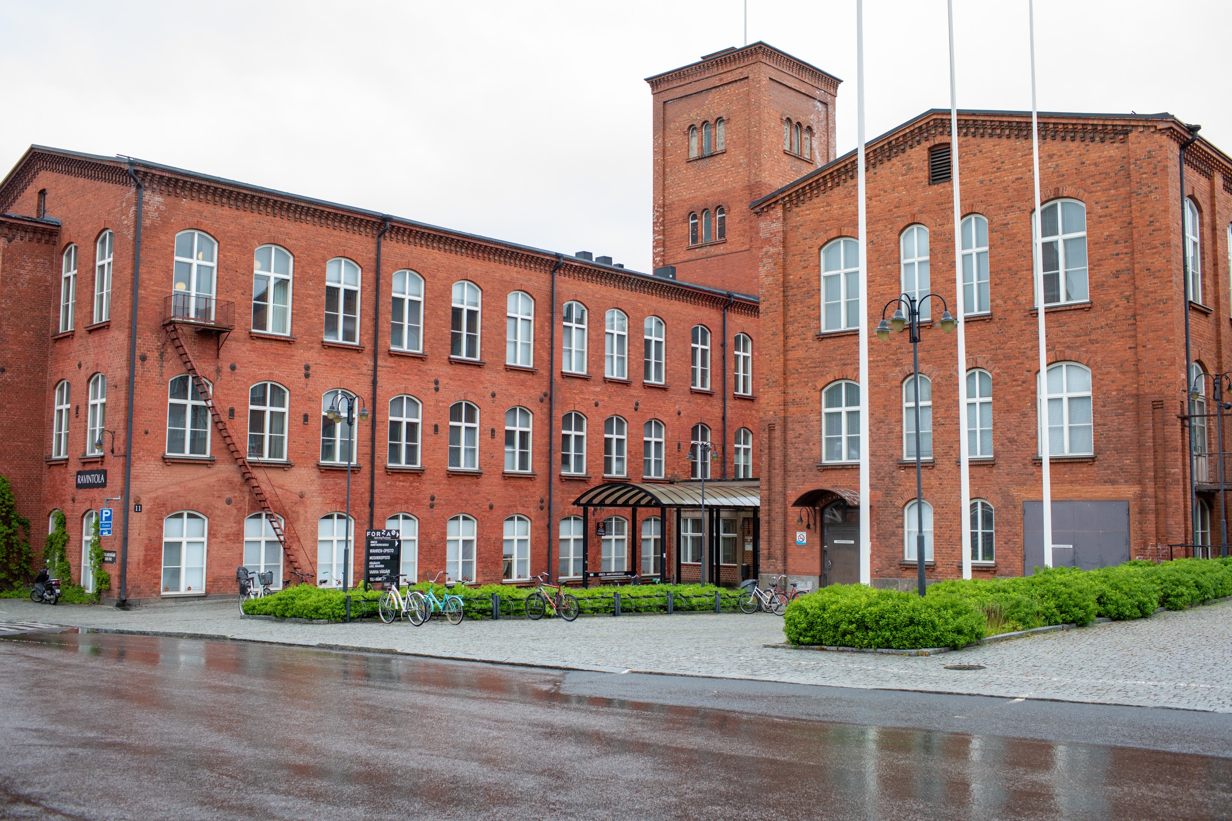 Forssa campus. Red brick, multi-storey building. Three flagpoles, bikes and green bushes in front of the building.