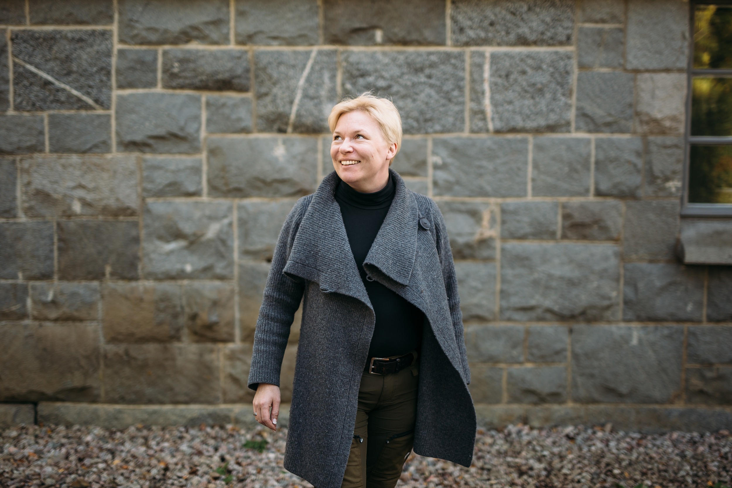 The person is standing in the middle of the picture, looking up at the distance with a smile. In the background is a wall of large stone blocks. 