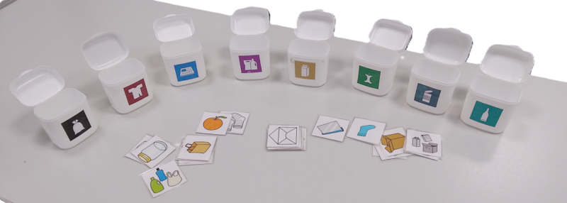 Kierrätetyistä ksylitolipastillipakkauksista tehdyt, jätepiktogrammeilla merkityt lajittelupelin astiat ja paperilaput, joissa kuvia eri jätteistä.