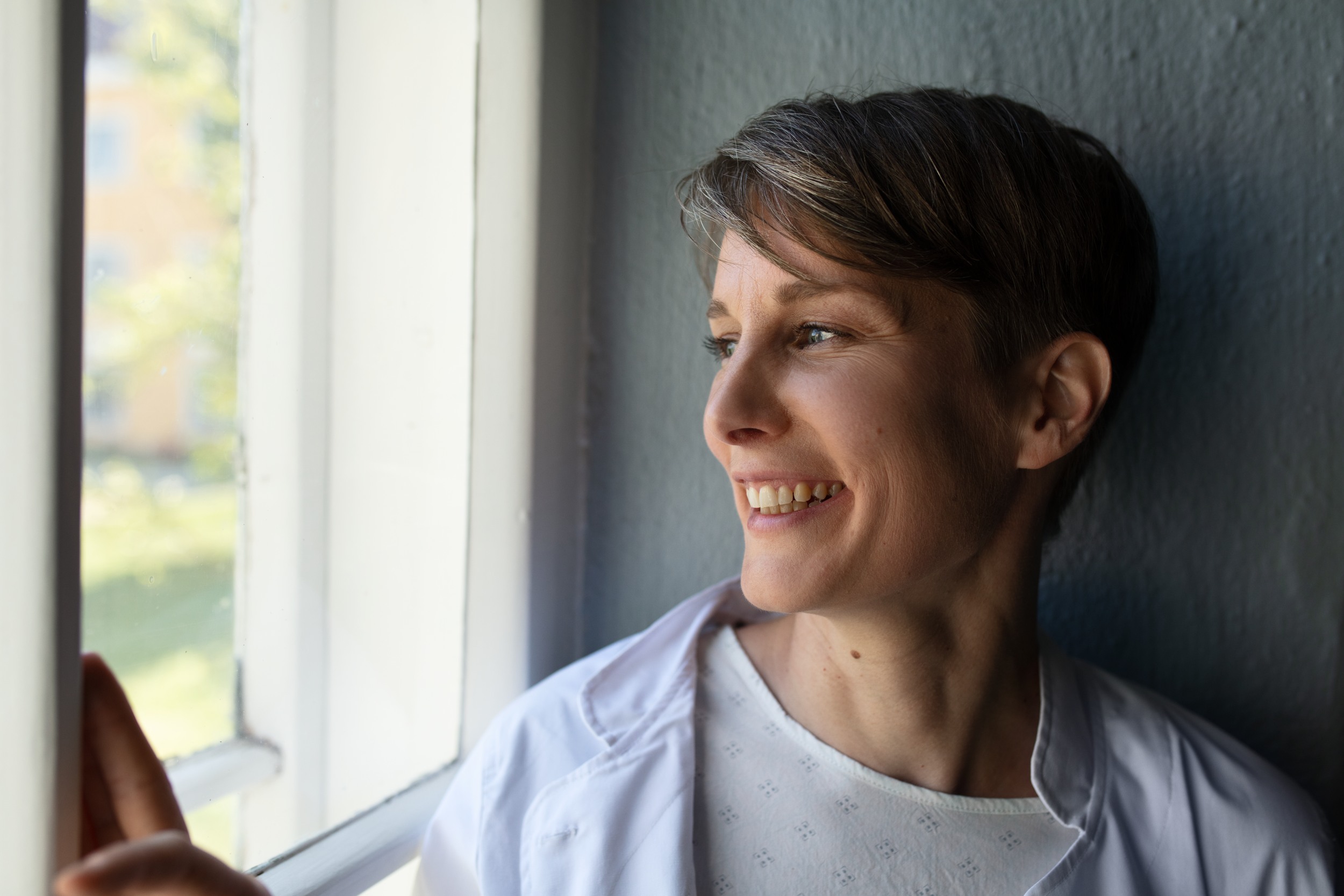 A person wearing a white jacket looks from window to outside and smiles.