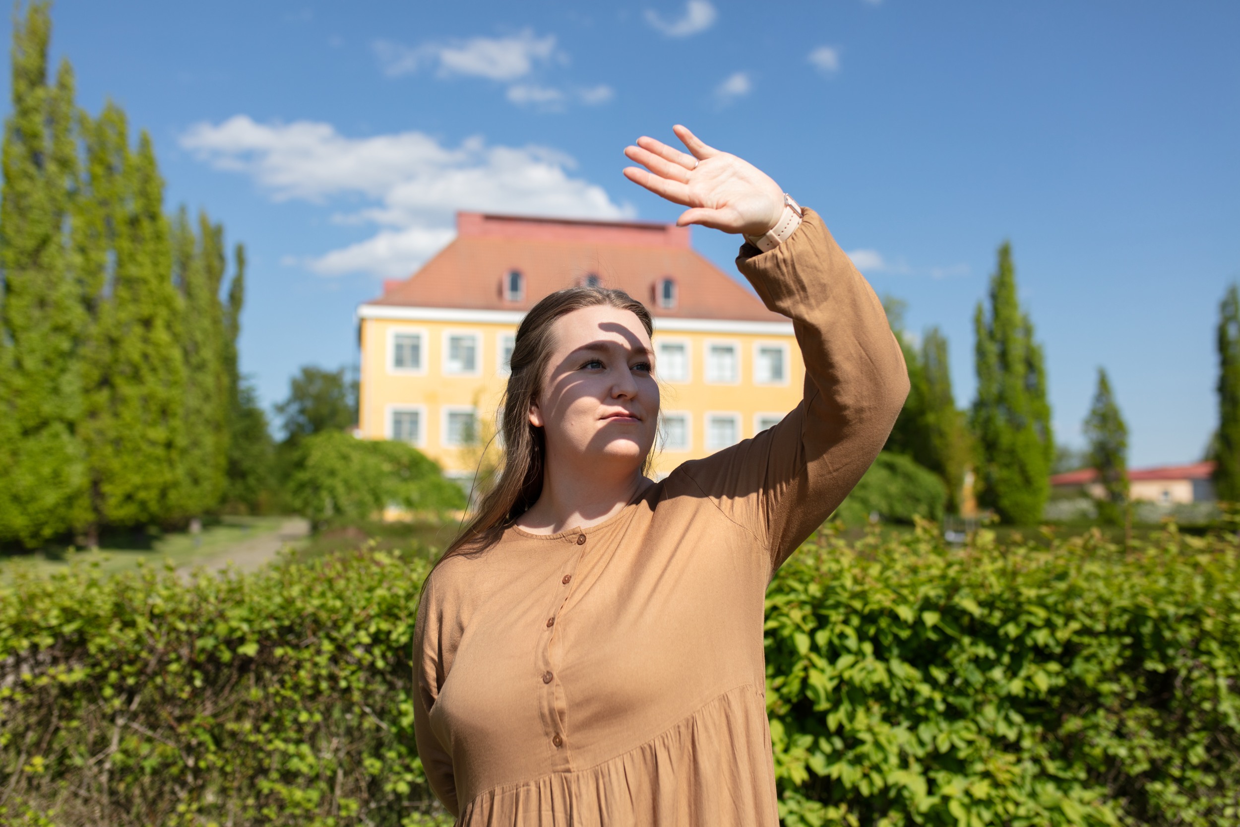 Nainen katsoo ylöspäin  ulkona ja varjostaa auringon heistumista silmiin kädellään. Takana keltaseinäinen ja punakattoinen vanha rakennus.