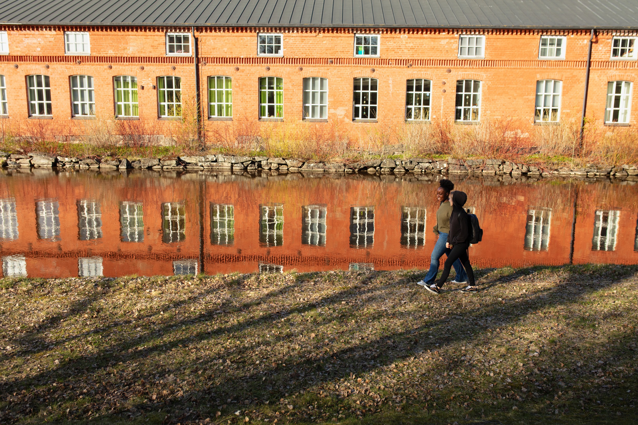 Two people walk along the riverbank, a yellow house reflected in orange in the water.
