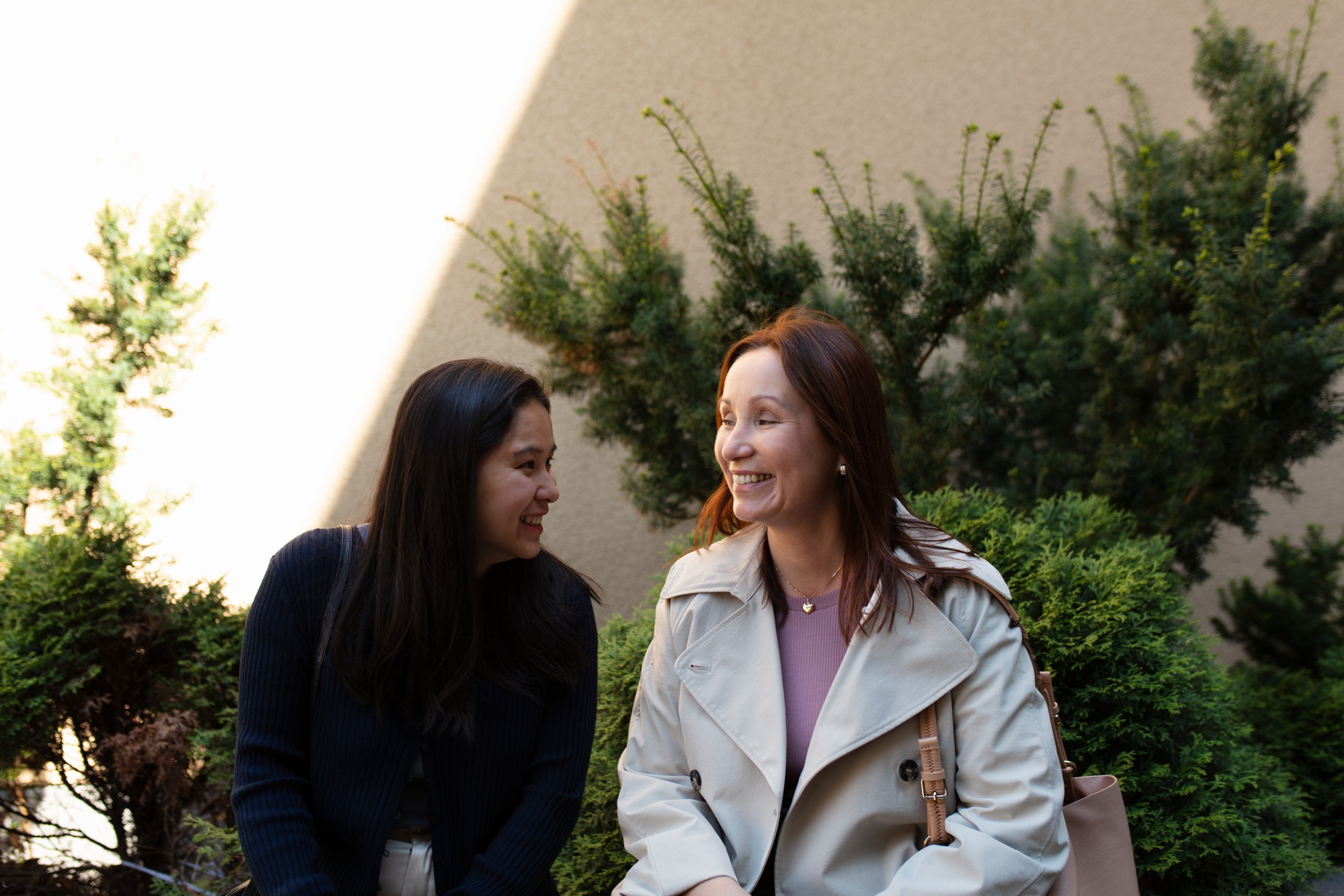 Two women are sitting in the shade of tree branches, laughing.