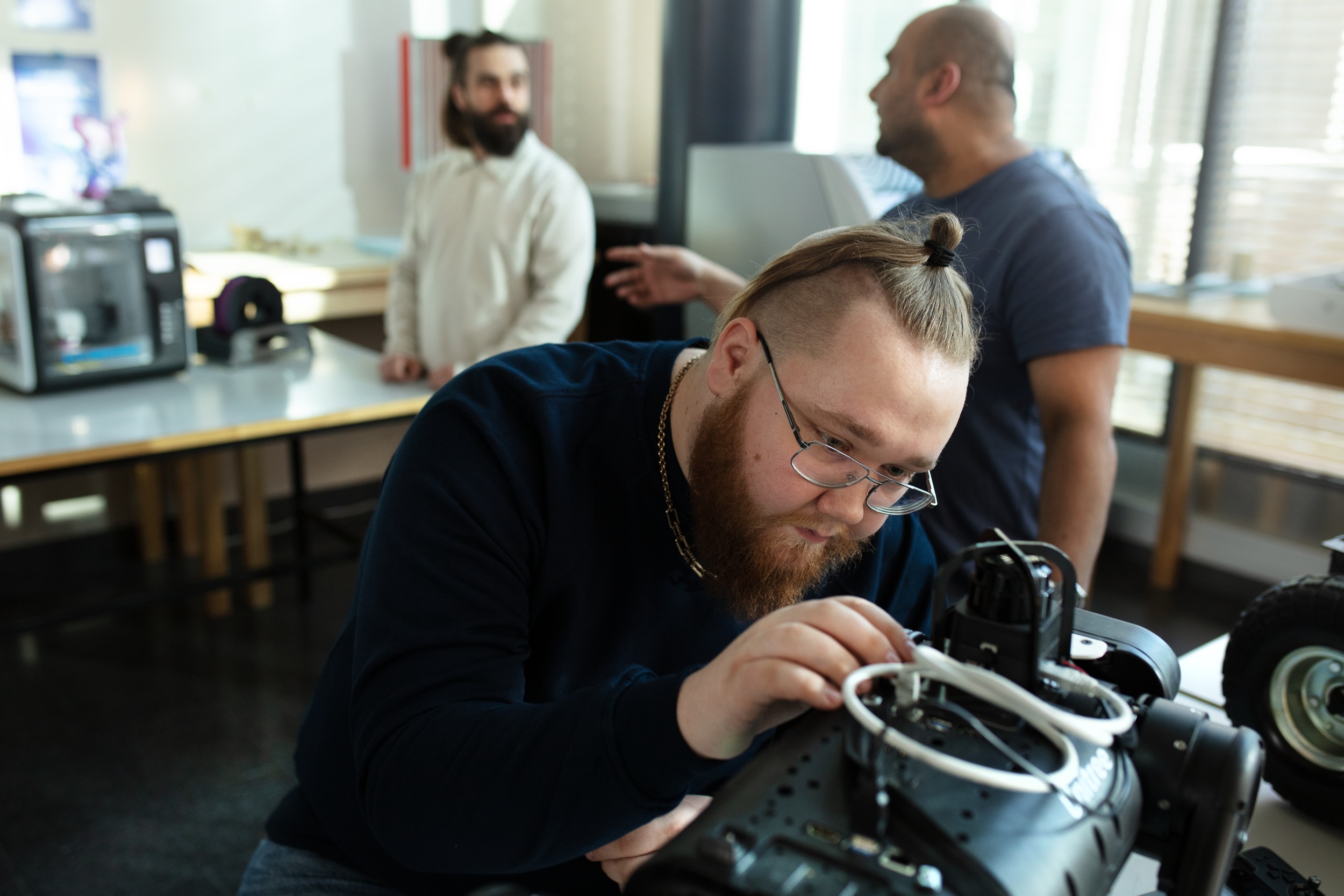 A person checks a machine. Two persons are discussing on the background.