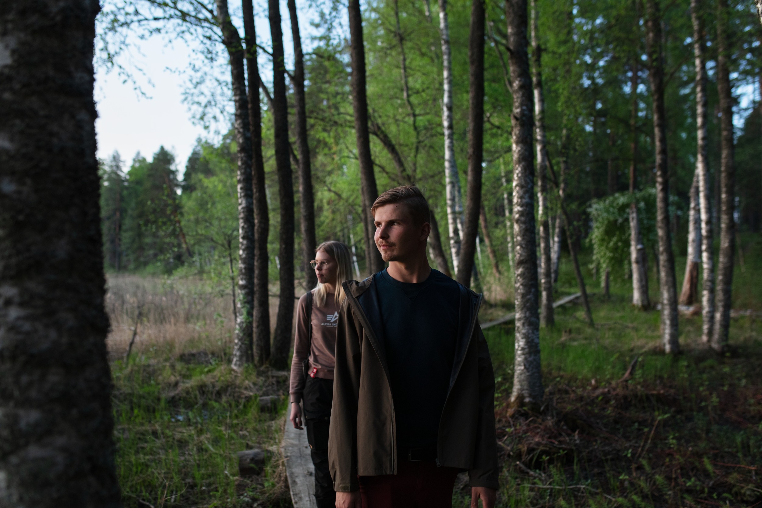 Two people walking on the forest trail. Young man in front and young woman behind, both looking left.