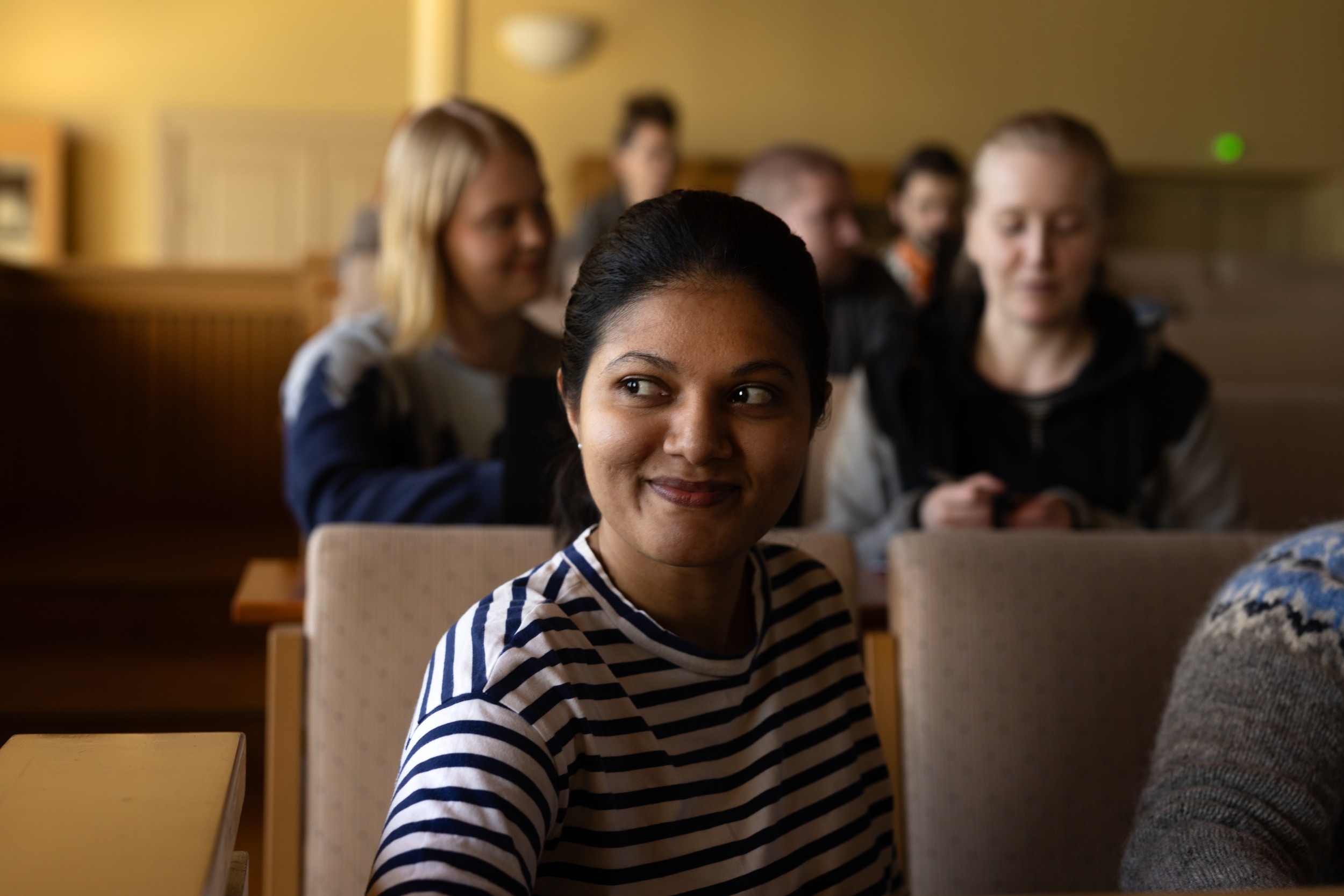 A dark-haired woman sitting in a classroom looks past the camera and smiles. behind other people sitting.