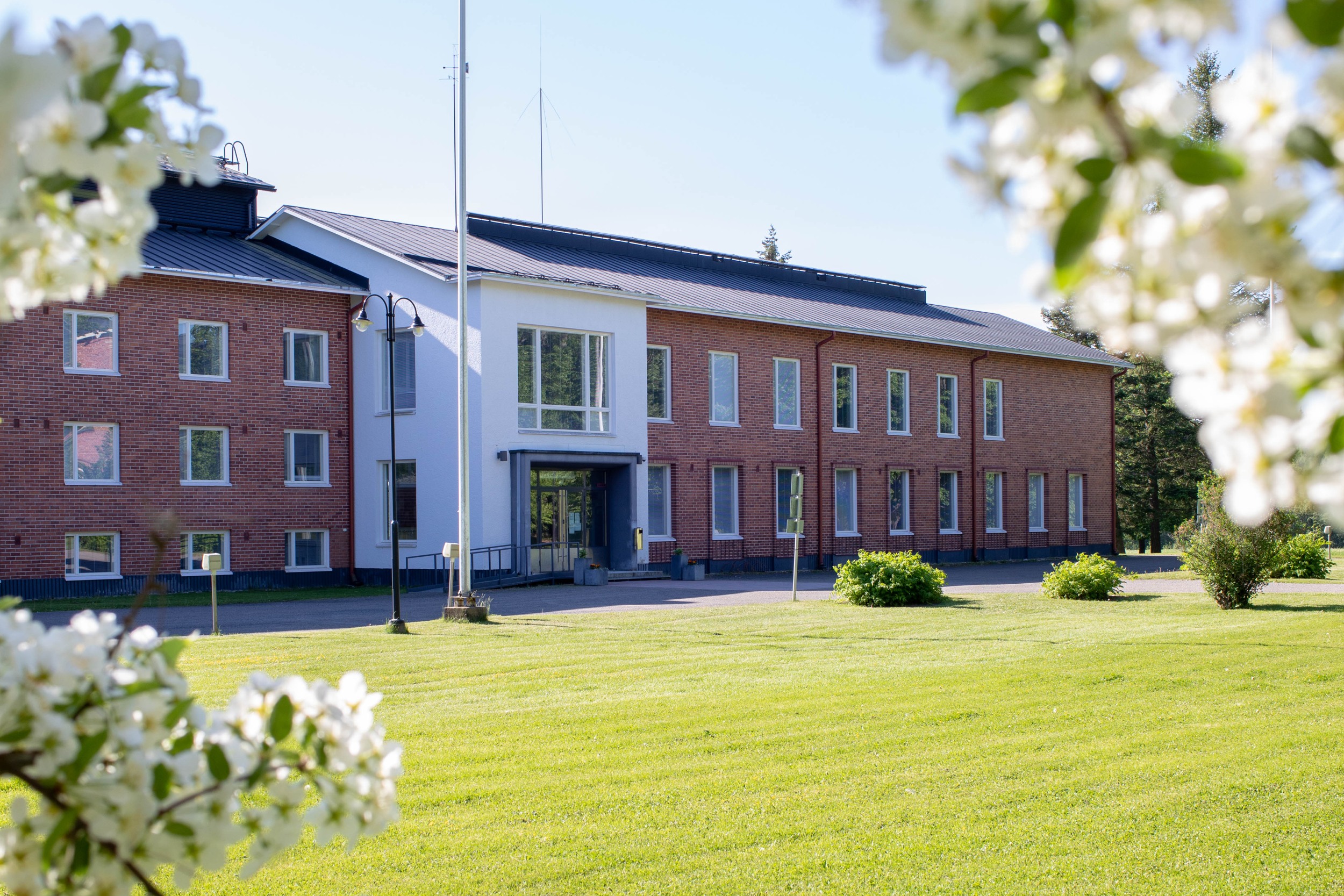The main building of the Mustiala campus. Red brick two-storey building. Lawn and flowering trees in the foreground.