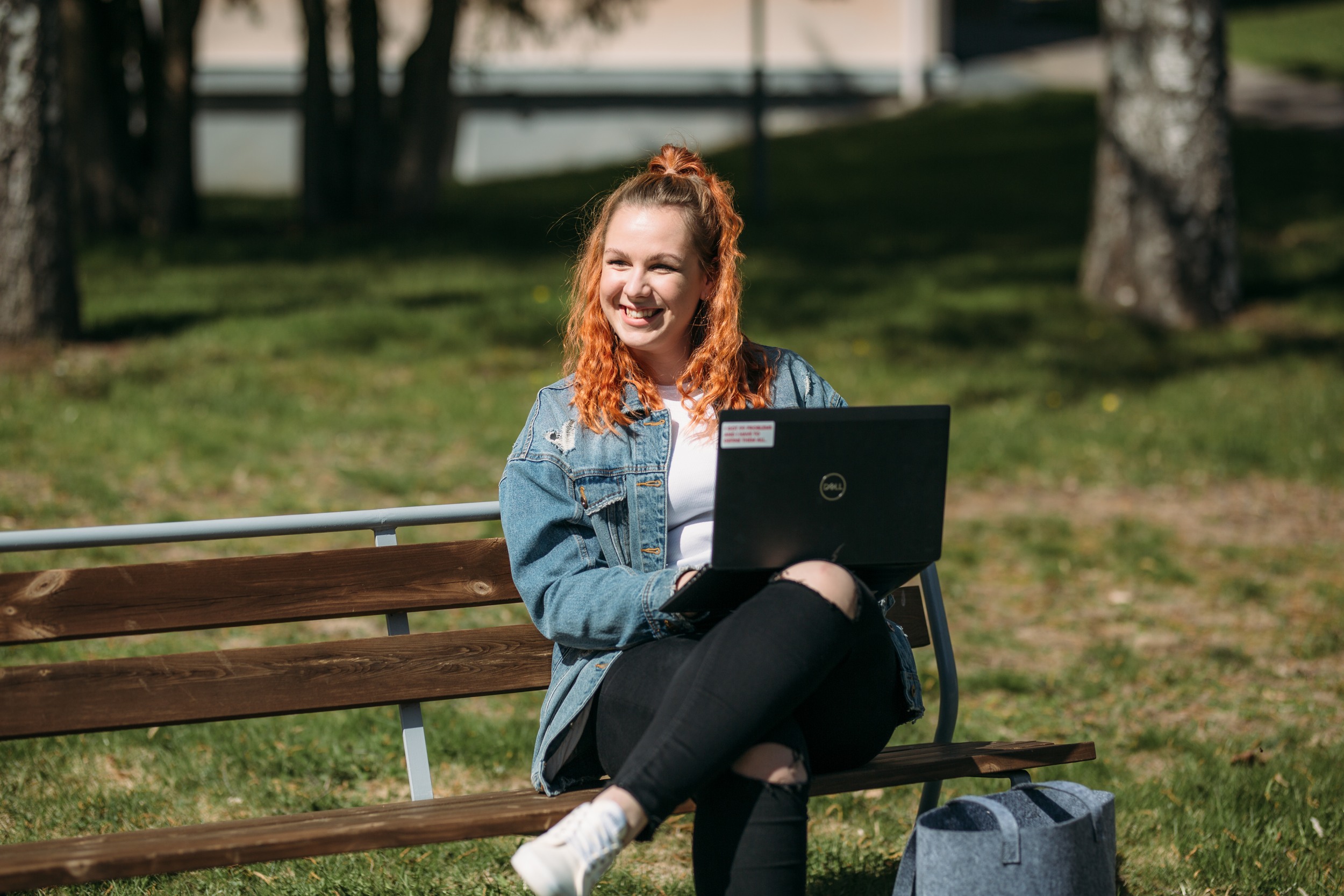 A red-haired woman sits on a park bench with a computer, smiling and looking past the camera.