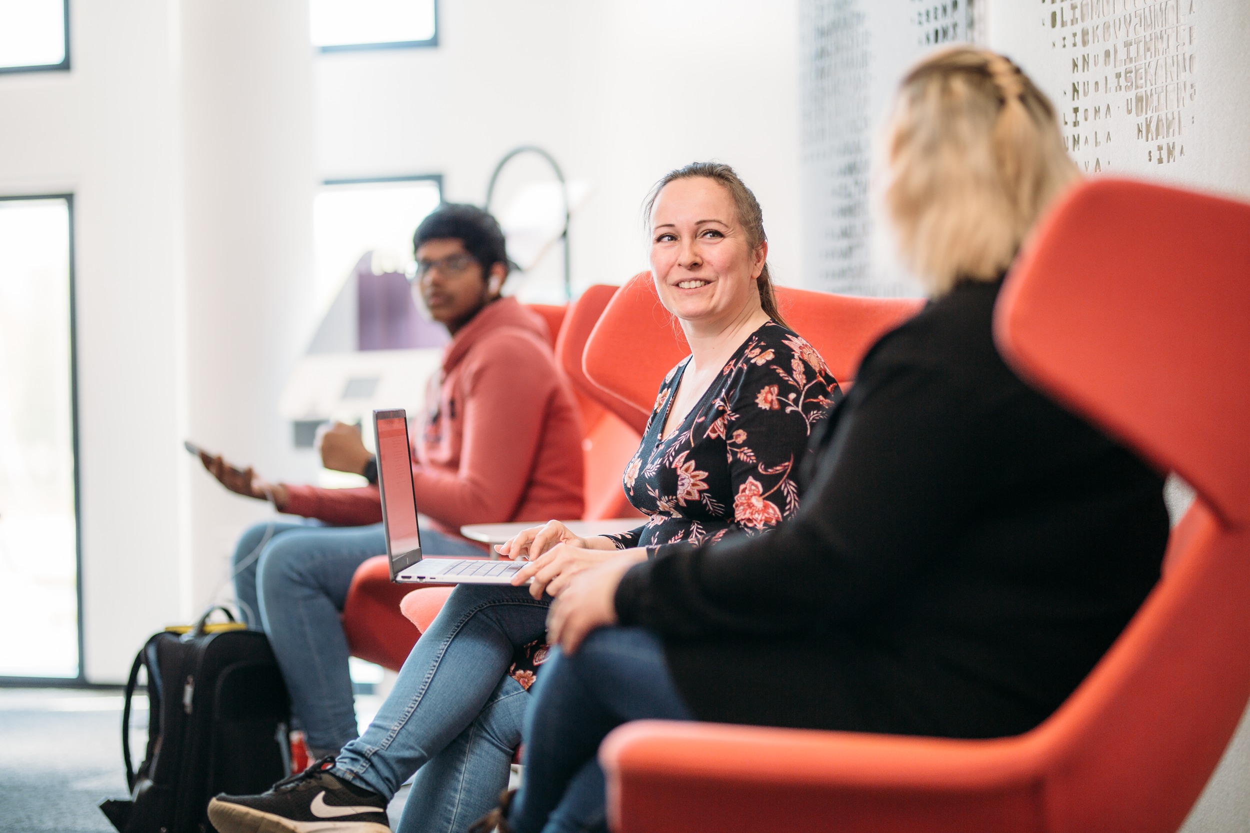 Three people are sitting on red chairs and talking. The middle person is clearly visible, the other two are blurred. In the background, a view of the University Centre.