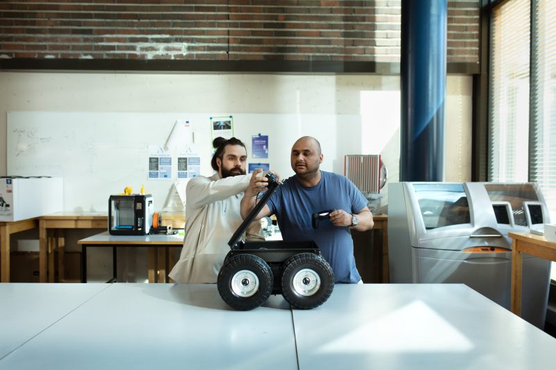 Two persons working with a robot, which has wheels and is on a table.