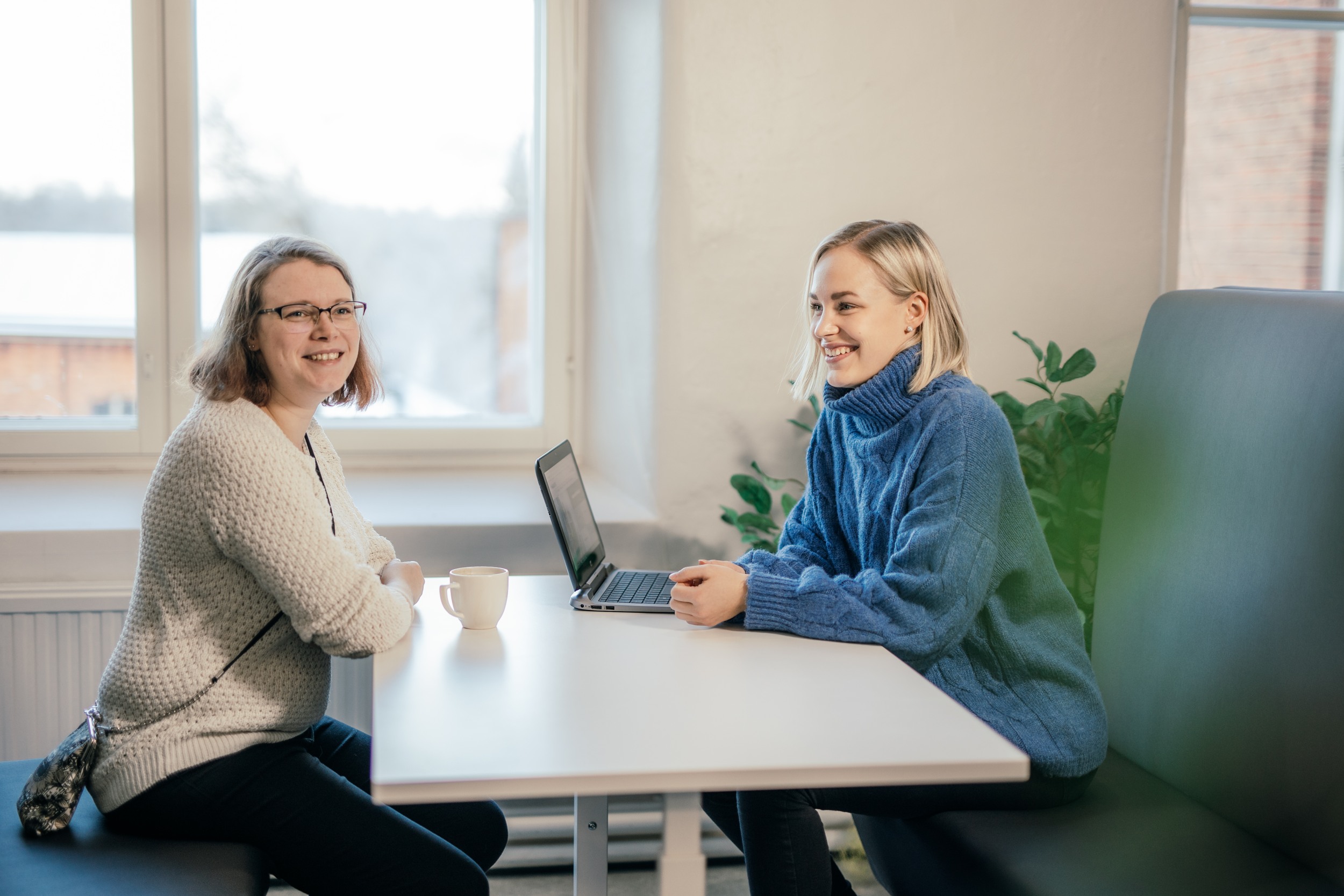 Kaksi henkilöä istuu pöydän ääressä ja hymyilee. Toisen henkilön vieressä pöydällä on kannettava tietokone. Taustalla näkyy talvinen maisema ikkunasta. 