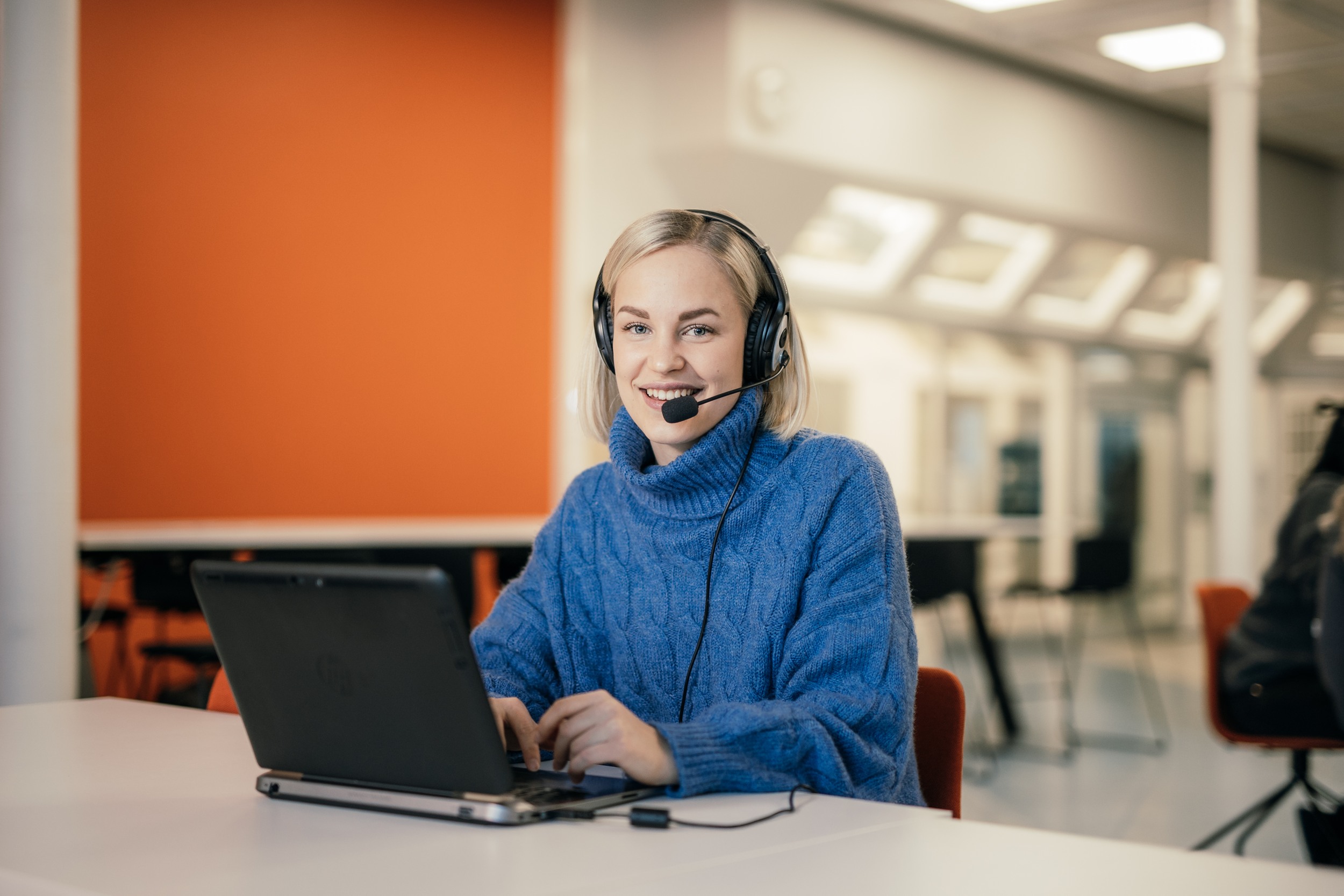 The person is sitting at a table, smiling towards the camera. On the table in front of him is a laptop and the person is wearing headphones with a microphone.