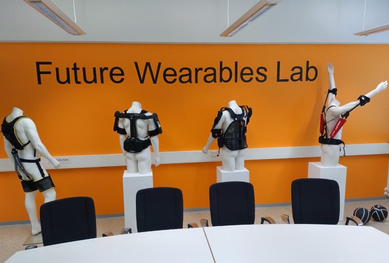 Neljä erilaista eksoskeletonia mallinukkejen päällä. Taustalla oranssi seinä, jossa lukee Future Wearables Lab