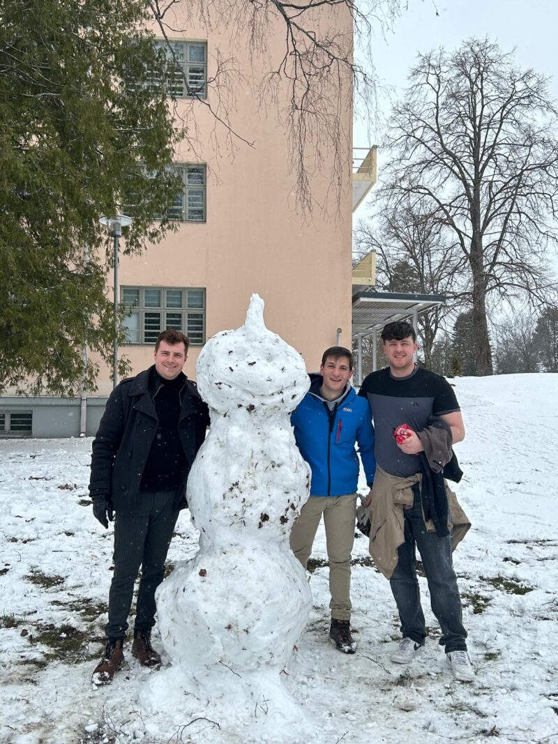 Three students standing outside next to a snowman