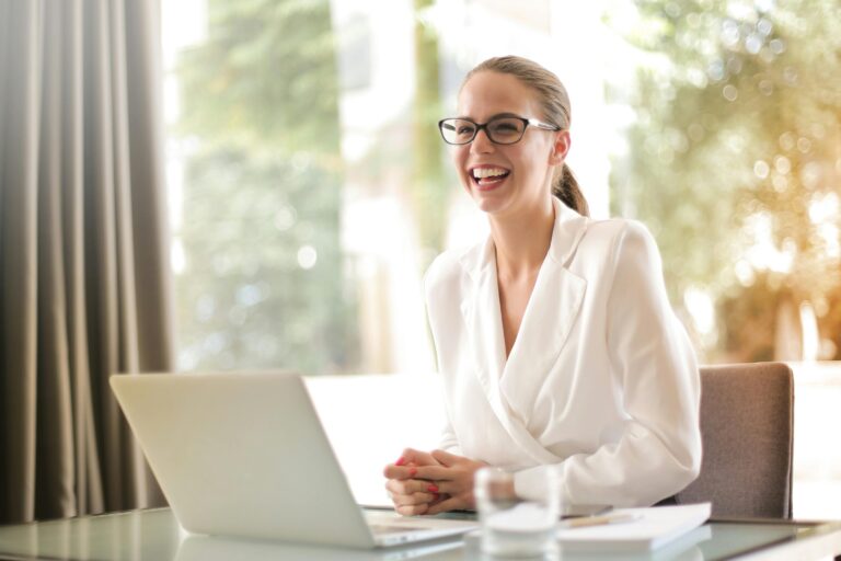 Light haired woman is sitting behind table and smiling with a laptop in front of her.