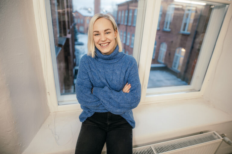 Nainen istuu hymyillen ikkunan edesssä/ Woman sitting in front of a window smiling