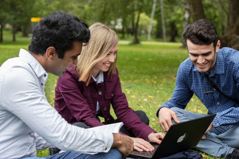 Kolme ihmistä istuu ulkona katsomassa tietokonetta/ Three people sitting outside looking at a computer