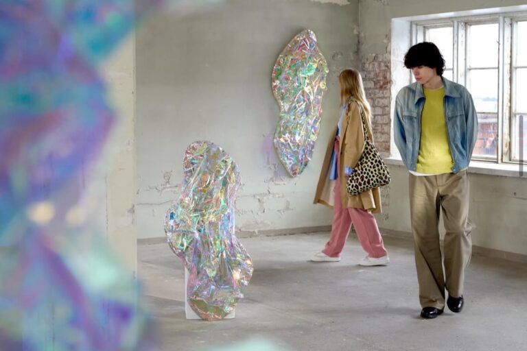 Two people walk through an exhibition space, where glass artworks are on display.