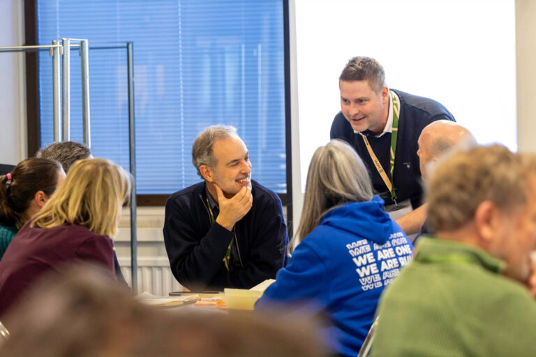 A group of adults engaged in a lively discussion around a table in a bright room during an event or workshop.