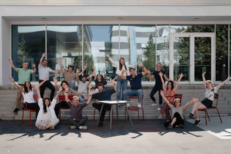 Lähes parikymmentä henkilöä ovat ulkona aurinkoisessa säässä, hymyilevät ja pitävät käsiään ylhäällä