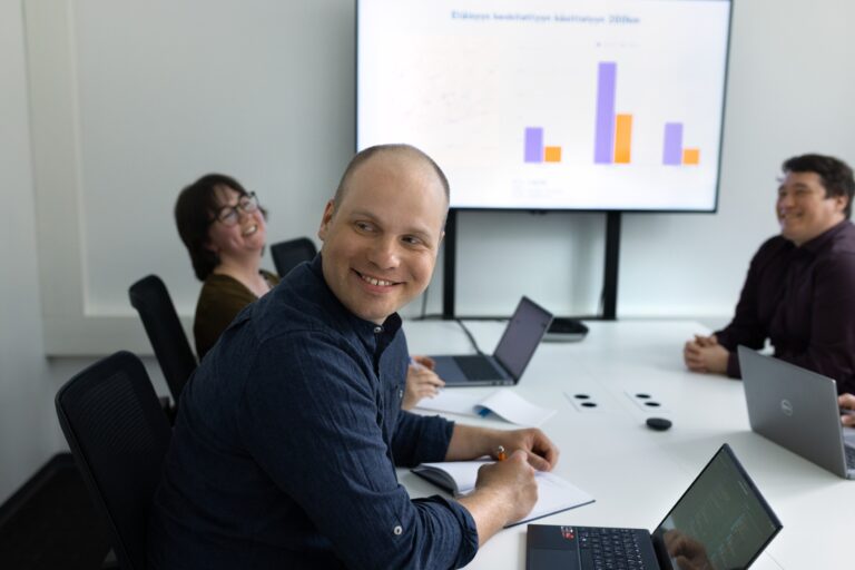 Kolme hymyilevää ihmistä istuu valkoisen neuvottelupöydän ympärillä kannettavat tietokoneet edessään- Seinällä oleva näyttö esittää värillisen pylväsdiagrammin.