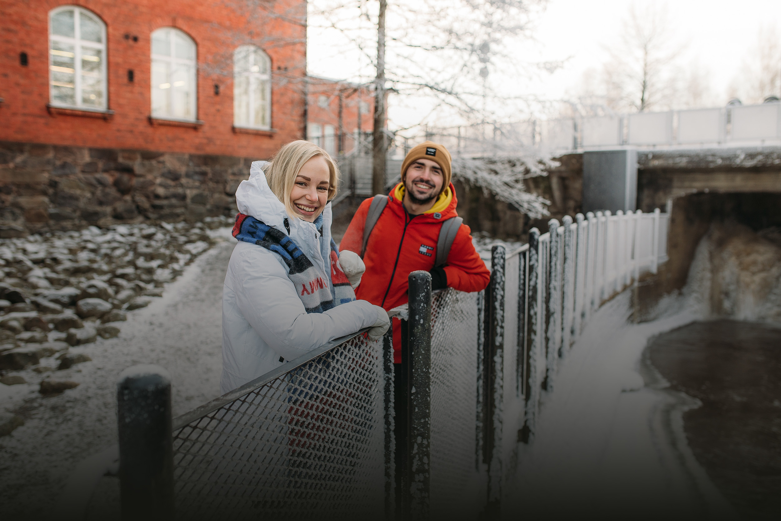 Kaksi henkilöä nojailee verkkoaitaan Forssan kampusmaisemassa talvisessa säässä.