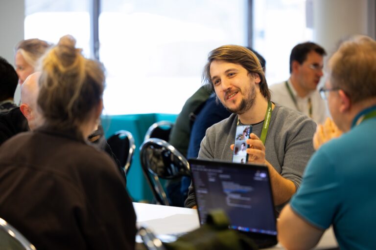 Ryhmä ihmisiä pöydän ääressä. Henkilö näyttää puhelimeltaan kuvaa muille osallistujille. Kuvituskuva.