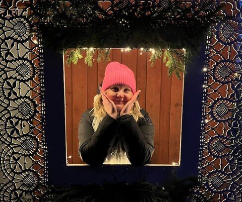 Iloiseva vaaleahiuksinen nainen pinkissä pipossa kurkistaa valokoristein ja koristekuviolla reunustetusta ikkunasta, kädet poskien alla hymyillen.