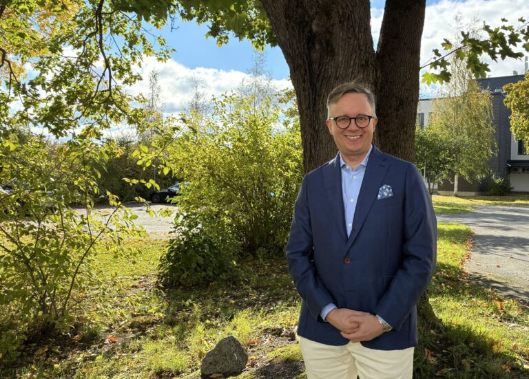 Sami Löfgren hymyilee puun varjossa kesäisessä maisemassa.