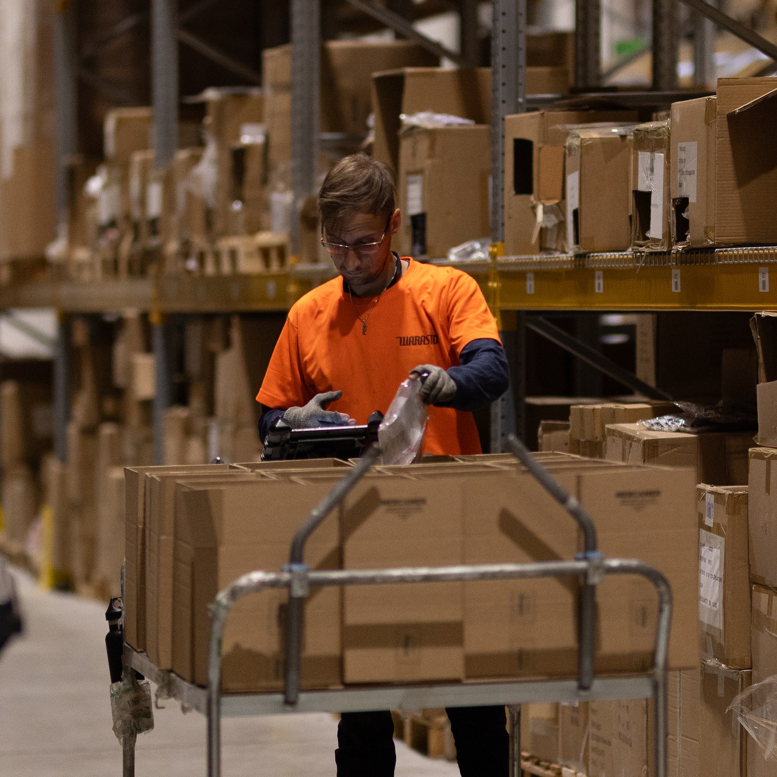 Varasto, jossa kirkkaan oranssissa paidassa oleva henkilö pallaa pahvilaatikoita.