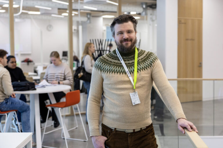 Mies seisoo käytävässä, jossa takana ihmisiä istumassa pöydän ääressä.