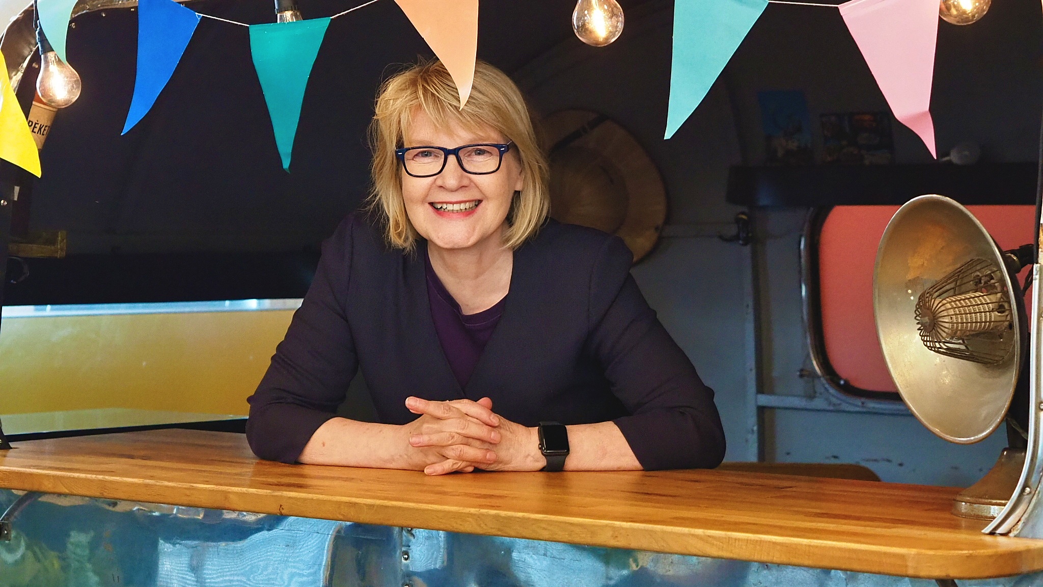 Hymyilevä henkilö nojaa puiseen tiskiin juhlavasti koristellussa tilassa. Taustalla roikkuu värikkäitä viirejä.