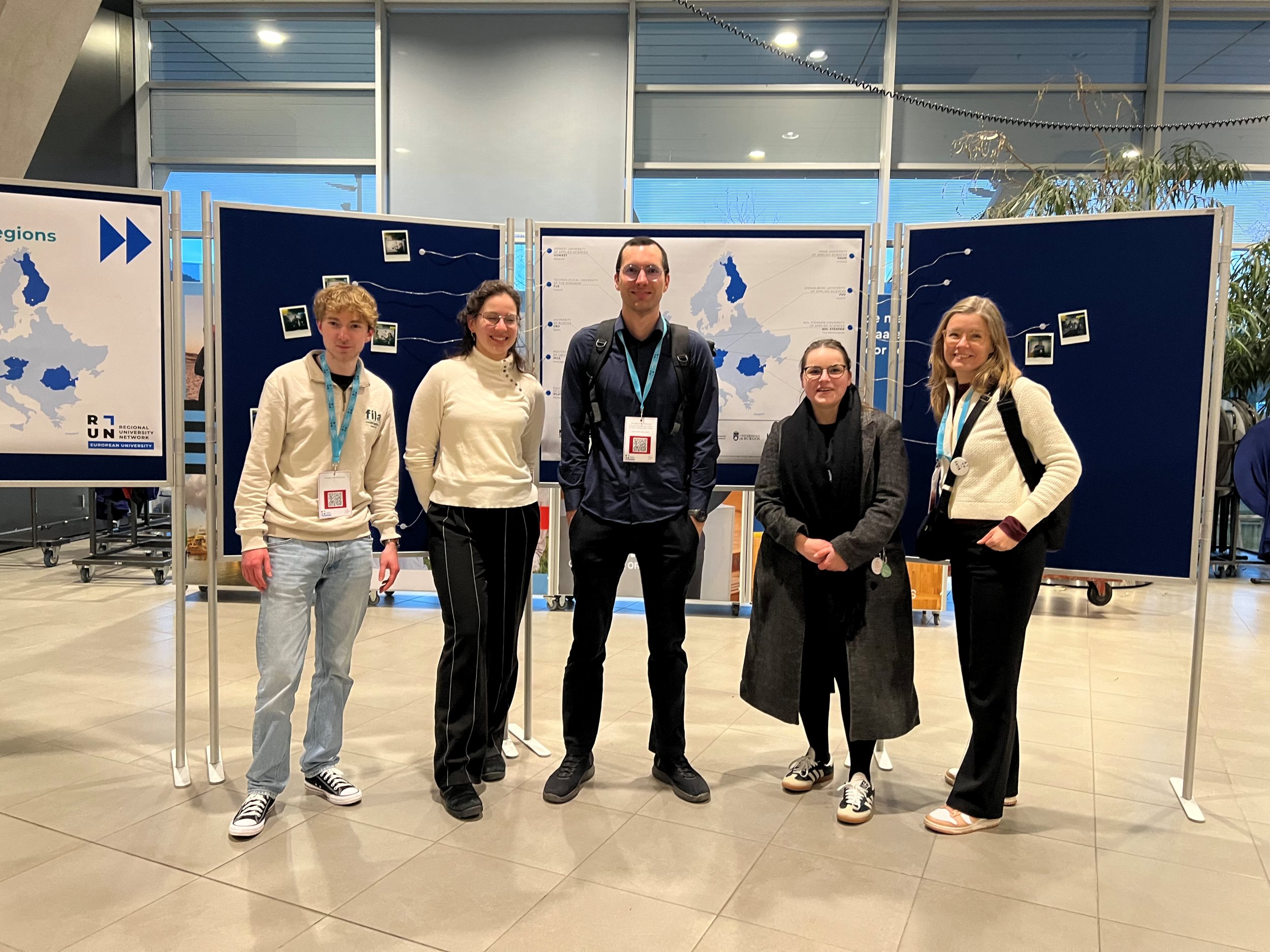 Viisi henkilöä seisoo vierekkäin sisätiloissa. Henkilöt katsovat kameraan ja hymyilevät