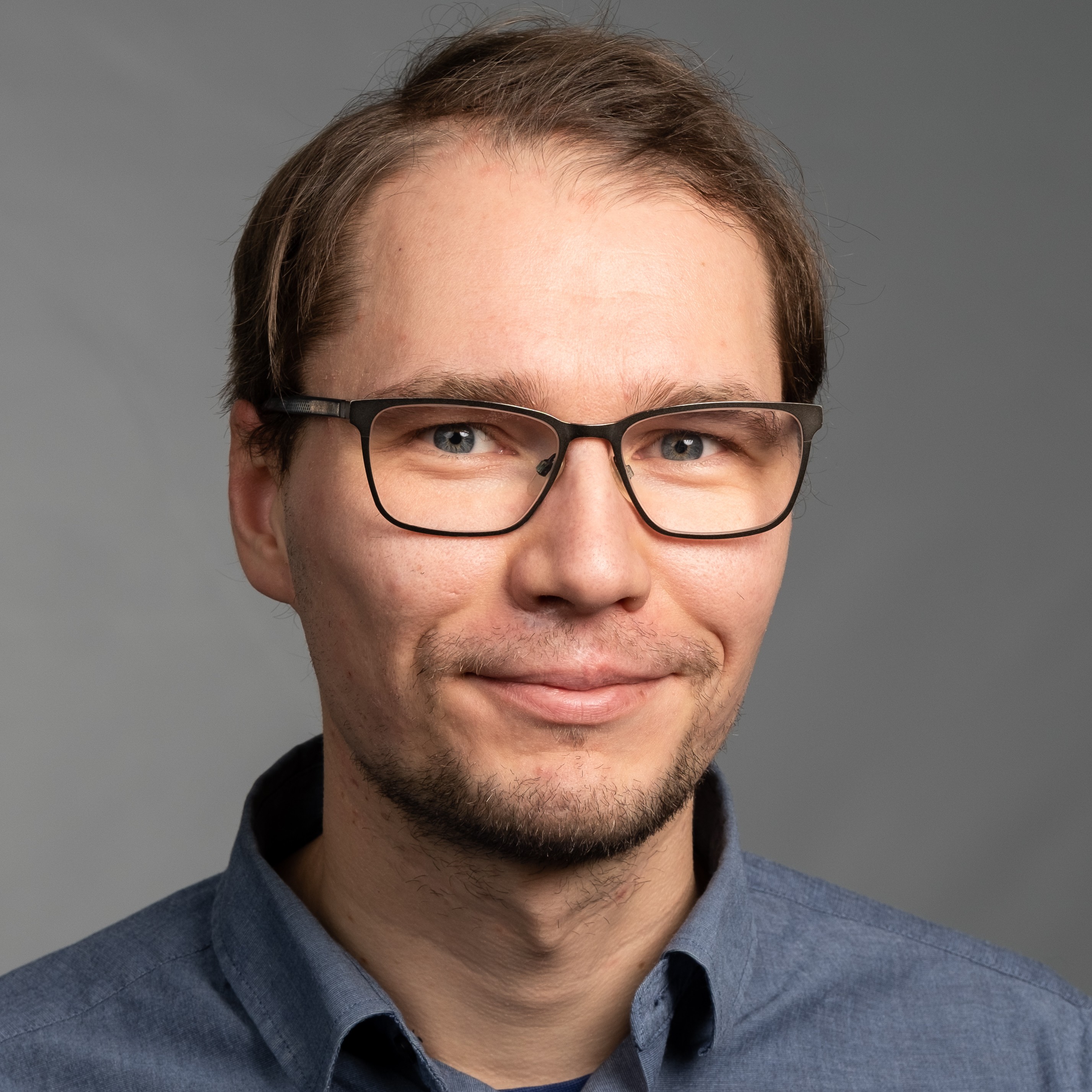 Henkilö, jolla silmälasit ja harmaa kauluspaita, hymyilee kameraa kohti.