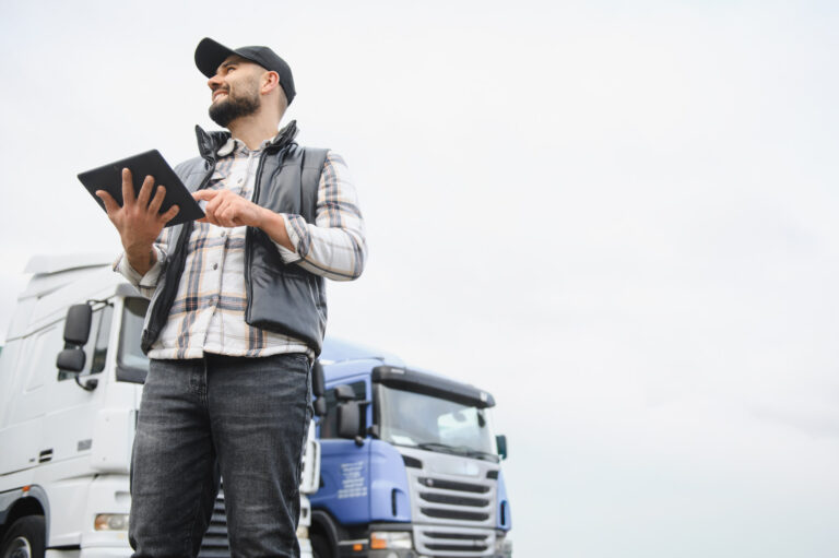 The driver is standing outside holding a tablet and looking to the side, with parked trucks and a cloudy sky in the background