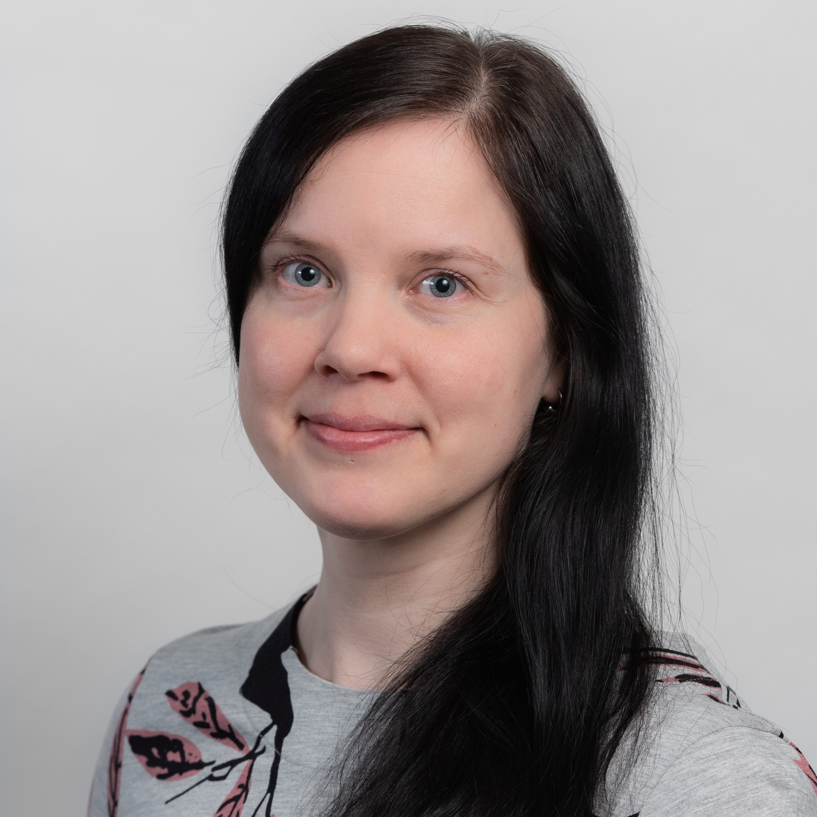 Henkilö, jolla tummat pitkät hiukset ja kukallinen paita. Katsoo kameraan.