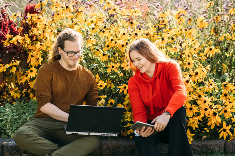 Two people outside with their laptops/ Kaksi ihmistä tietokoneilla puutarhassa
