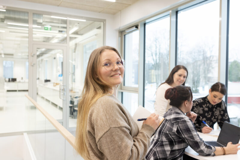Henkilö katsoo kameraan. Kolme henkilöä taustalla tekevät ryhmätyötä