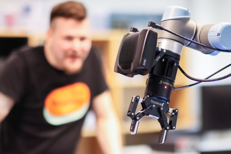 Lähikuva robottivarresta, jonka päähän on kiinnitetty kamera ja puristin, taustalla sumeana henkilö seuraamassa robotin toimintaa.