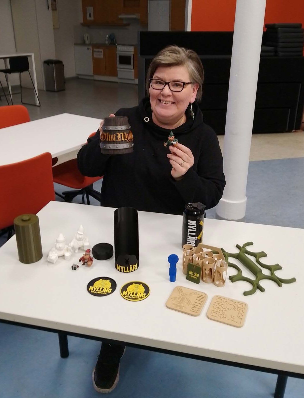 Henkilö esittelee OlutMyllylle suunniteltuja 3D-tulostettuja prototyyppejä. Pöydällä on muun muassa mukinalusia, olutkahvoja ja pieniä 3D-hahmoja.