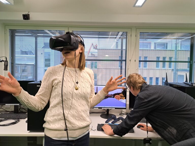 Tietokoneluokka, etualalla VR‑lasit päässään seisova henkilö kädet levitettyinä kokeilemassa virtuaalitodellisuutta, taustalla toinen henkilö kumartuneena säätää tietokonetta ikkunasta näkyy rakennuksia