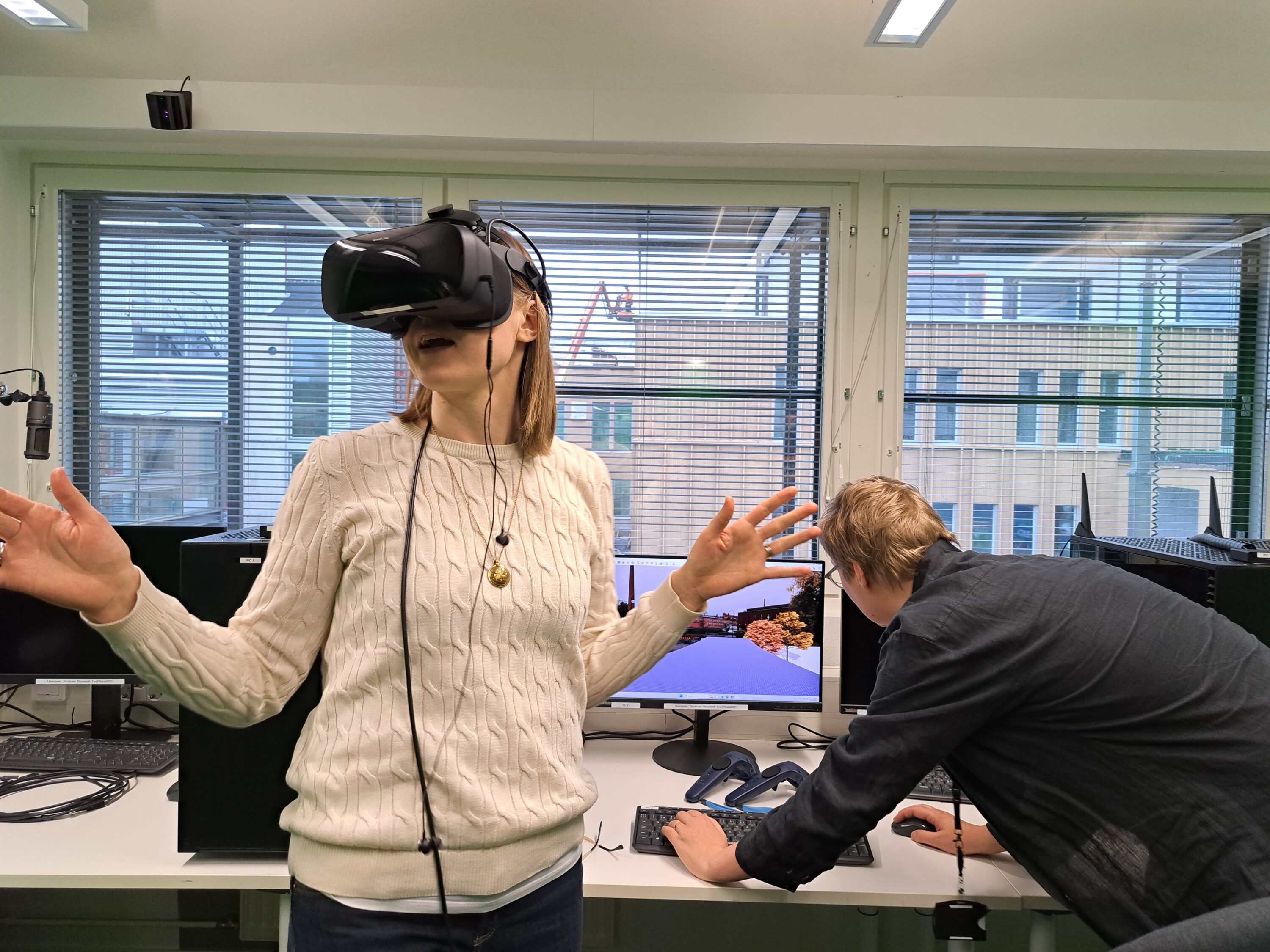 Tietokoneluokka, etualalla VR‑lasit päässään seisova henkilö kädet levitettyinä kokeilemassa virtuaalitodellisuutta, taustalla toinen henkilö kumartuneena säätää tietokonetta ikkunasta näkyy rakennuksia