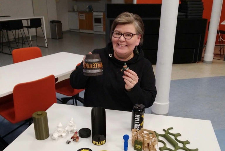 Henkilö esittelee OlutMyllylle suunniteltuja 3D-tulostettuja prototyyppejä. Pöydällä on muun muassa mukinalusia, olutkahvoja ja pieniä 3D-hahmoja.