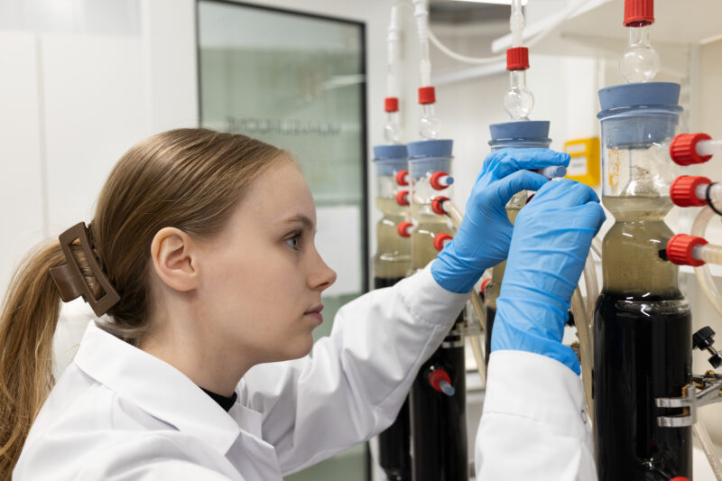Henkilö valkoisessa laboratoriotakissa ja sinisissä suojakäsineissä säätää lasisia kolonneja ja putkistoa laboratoriotyötilassa.