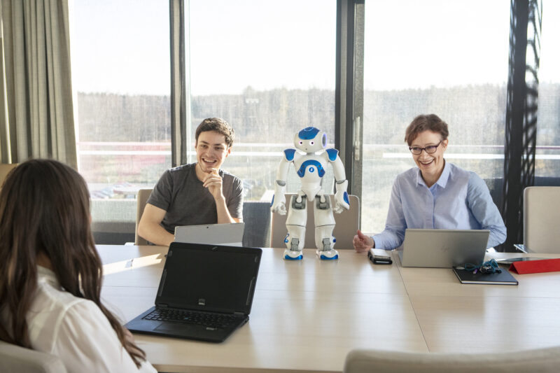 Three people sitting in a classroom with a robot/Kolme ihmistä istuu luokassa robootin kanssa. 